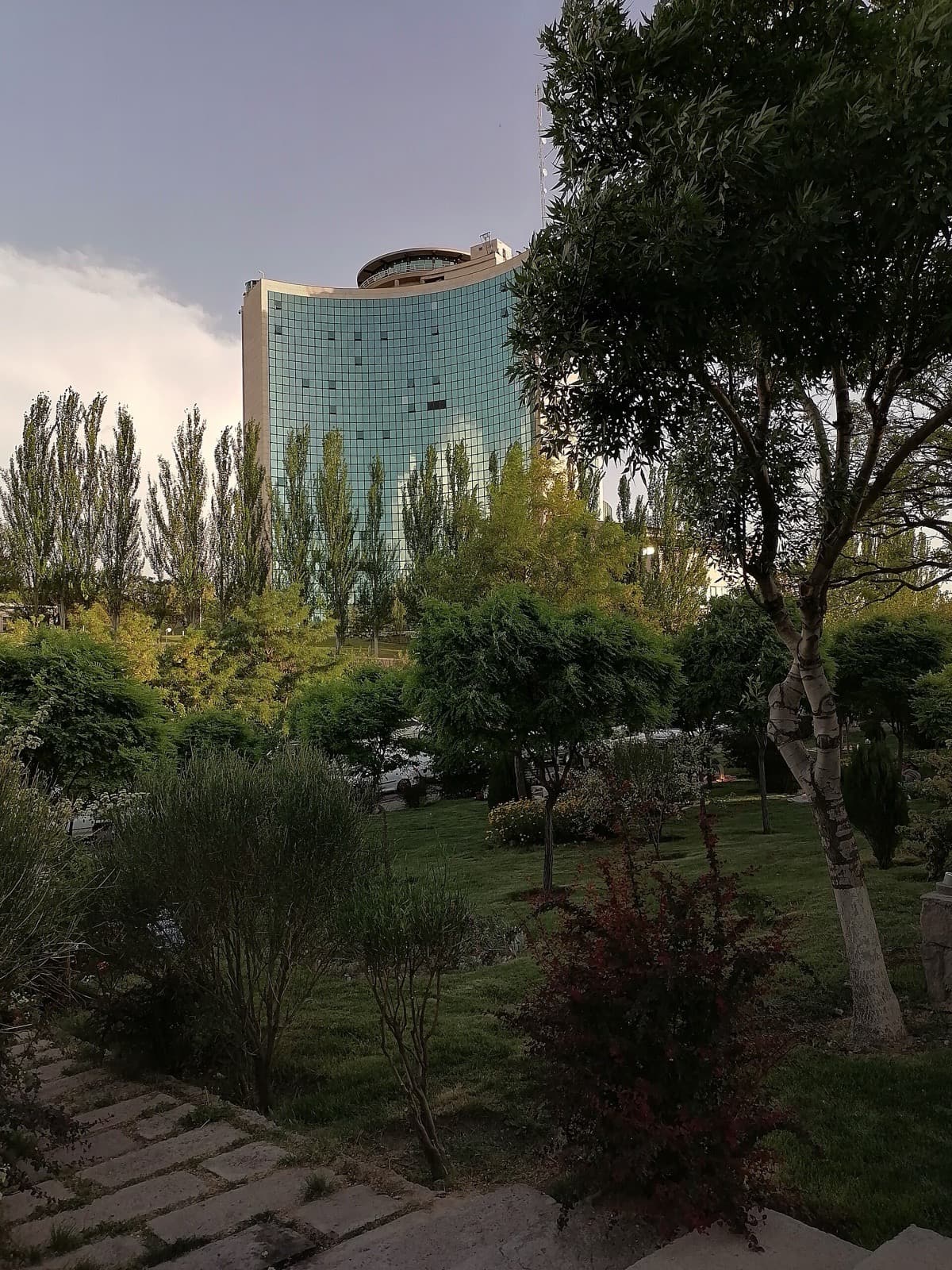 برج منحنی شیشه‌ای هتل پارس ائل‌گلی دیده‌شده از میان درختان پارک